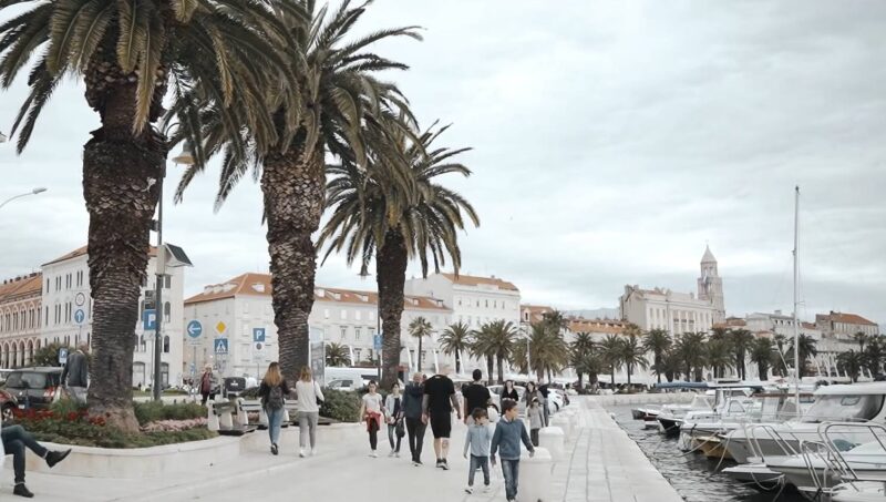 Ljudi šetaju uz obalu Splita pored palmi i usidrenih brodova